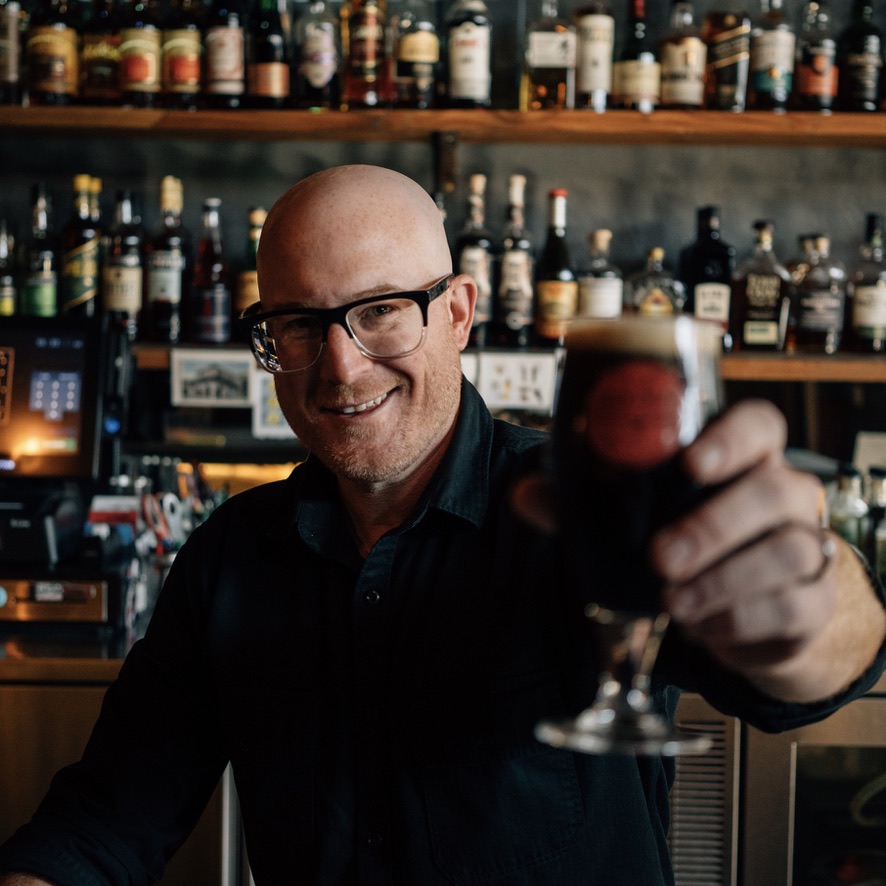 Chris Cohen, Advanced Cicerone, raising a glass of dark beer behind the bar
